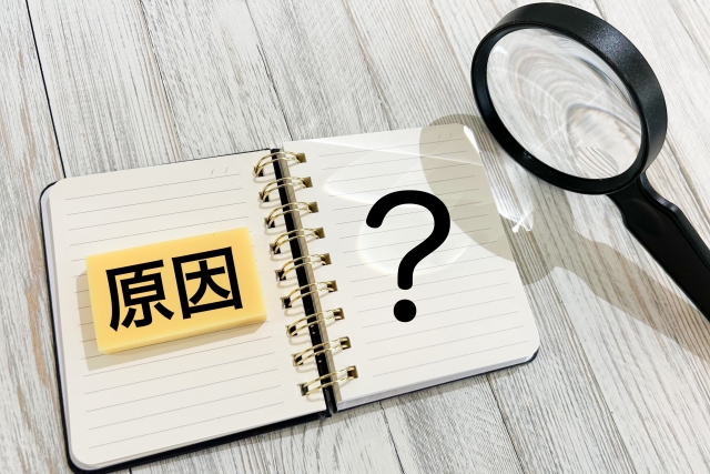 ノートに「原因？」と書かれ、虫眼鏡が置かれている写真。問題やトラブルの原因を調べるイメージを表現。