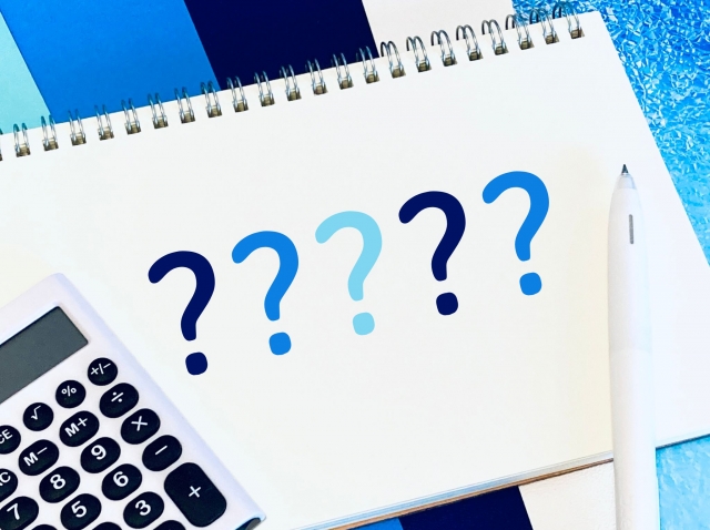 ノートに並ぶ疑問符と電卓、ペン、クリップボードが置かれた写真。買取サービスの選び方や比較検討をイメージした構図。